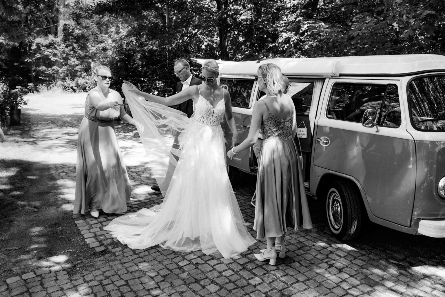 Braut steigt aus dem VW-Bully aus und richtet mit ihren Brautjungfern ihr Hochzeitskleid