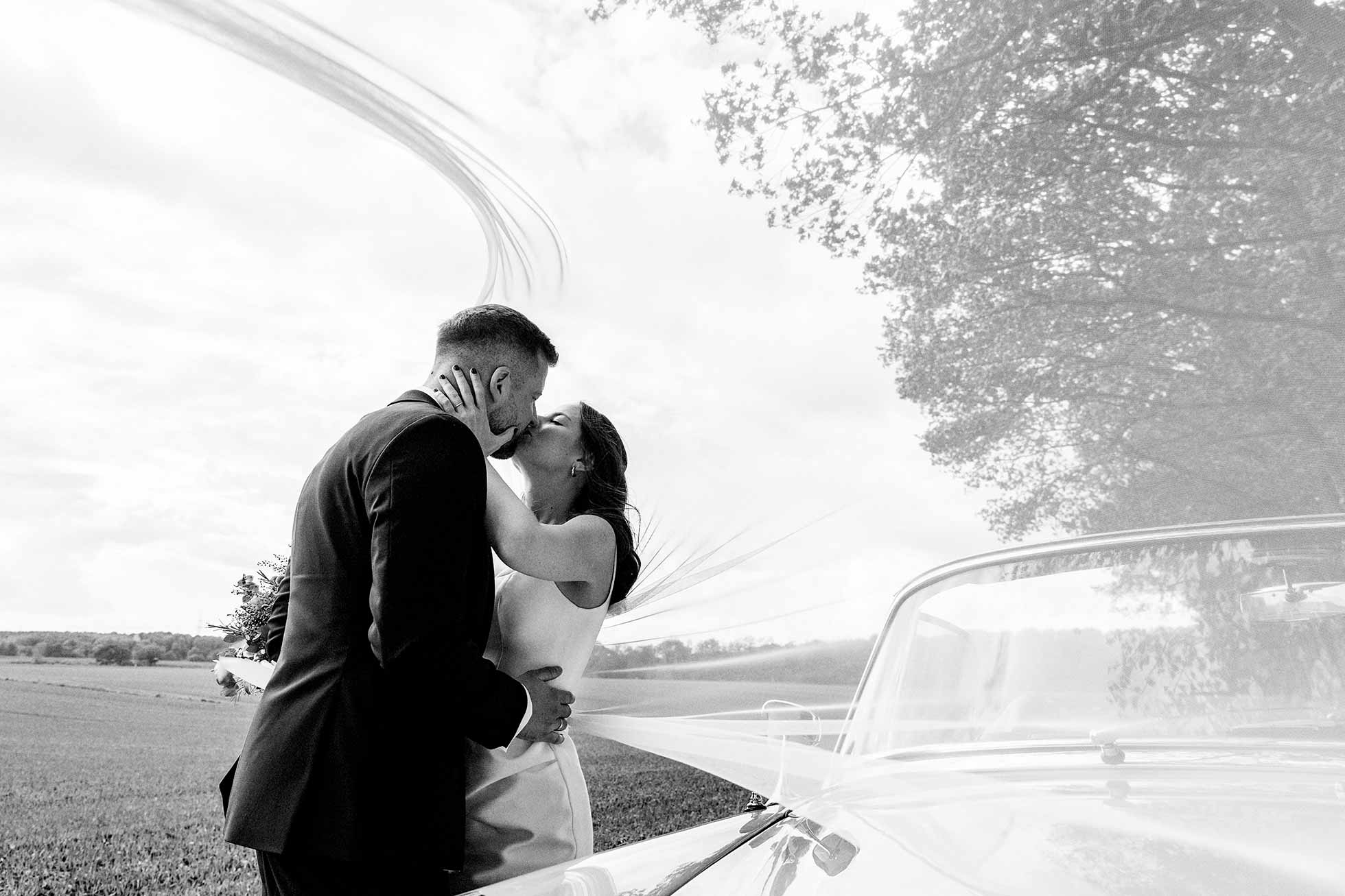 Ein küssendes Brautpaar unter einem Schleier. Im Vordergrund sieht man das Hochzeitsauto.