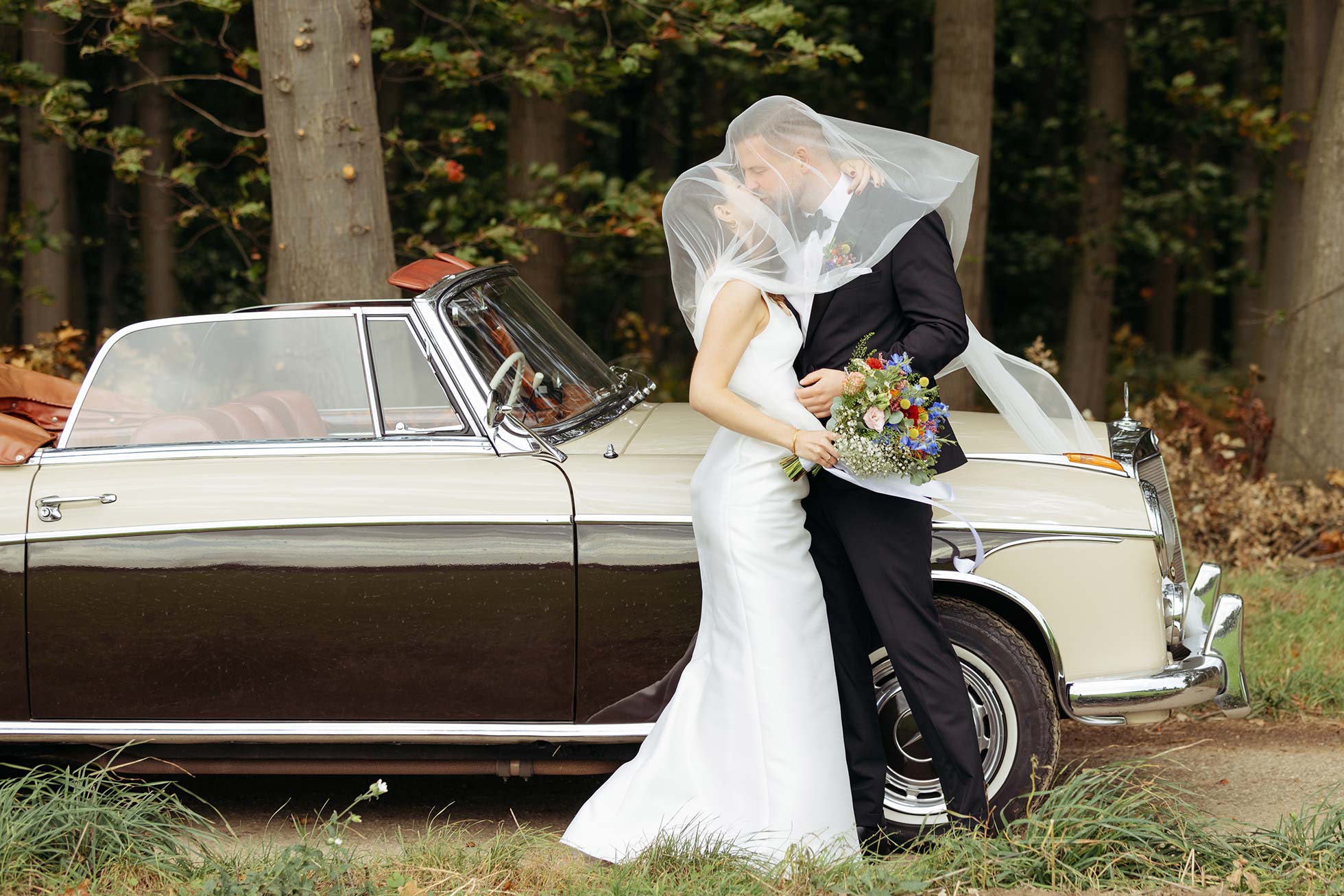 Ein küssendes Brautpaar vor dem Hochzeitauto. Der Schleier der Braut wäht über die Köpfe des Paares.