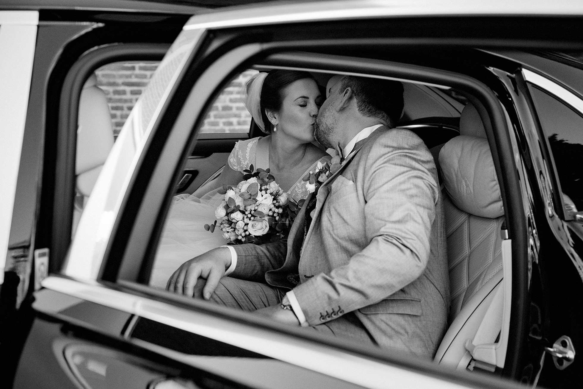 Ein Brautpaar küsst sich im Hochzeitsauto. Dieses schwarzweiss Hochzeitsfoto wurde nach der kirchlichen Trauung in Neuss gemacht.